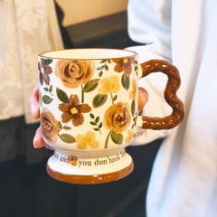 A hand holding a single vintage aesthetic cream ceramic pedestal mug with brown and yellow rose patterns, featuring a unique brown wavy handle and script text on its footed base, over a white surface.