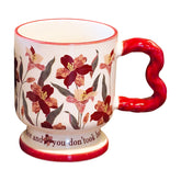 Isolated vintage aesthetic ceramic pedestal mug on a white background, featuring dark red floral oil painting patterns and a red wavy handle. Elegant script text is partially visible on the footed base.