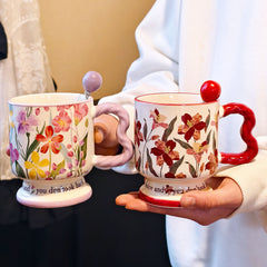 A hand holding two vintage aesthetic ceramic pedestal mugs side-by-side against a soft background. The left mug has purple and pink watercolor flowers with a lavender wavy handle; the right mug has dark red floral patterns with a red handle. Script text is visible on both bases.