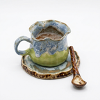 A product shot against a pure white background featuring a handmade, wabi-sabi style ceramic cup, saucer, and spoon set. The irregular-shaped cup has a wavy rim and features a distinctive glaze pattern of pale green on the lower body and a mottled blue-gray on the upper body and handle. It sits on a matching blue-gray and brown glazed saucer. To the right is a small ceramic spoon with a twisted brown handle.