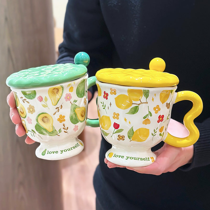 Two aesthetic pedestal mugs with textured ceramic lids and wavy handles, held by a person. The left mug features green avocado and floral patterns with a green handle and lid; the right mug displays lemon and yellow flower patterns with a yellow handle and lid. Both bases feature the text 'love yourself.'