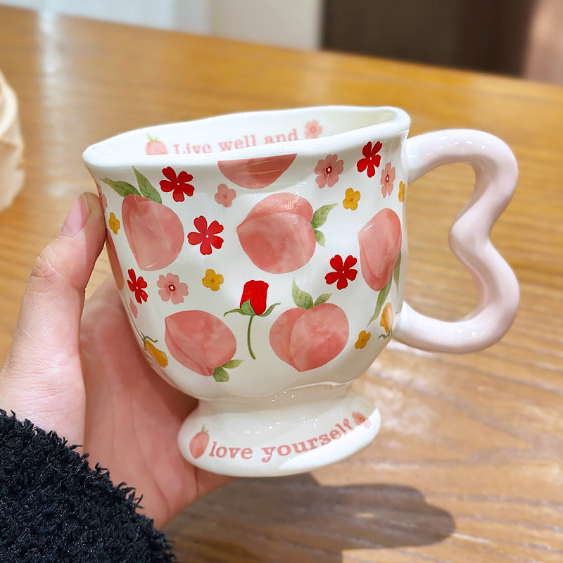 Aesthetic cream ceramic pedestal mug with sweet pink peach and red floral patterns, featuring a light pink wavy handle and the text 'love yourself' on its footed base, held by a hand.