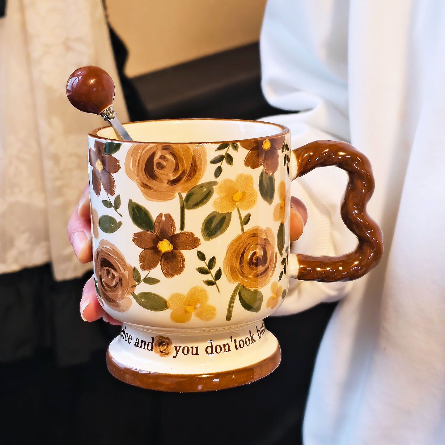 A hand holding a vintage aesthetic cream ceramic pedestal mug with brown and yellow rose patterns and a brown wavy handle, with a matching brown spherical ball ornament stirring spoon placed inside. Script text is visible on the base.