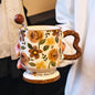 A hand holding a vintage aesthetic cream ceramic pedestal mug with brown and yellow rose patterns and a brown wavy handle, with a matching brown spherical ball ornament stirring spoon placed inside. Script text is visible on the base.