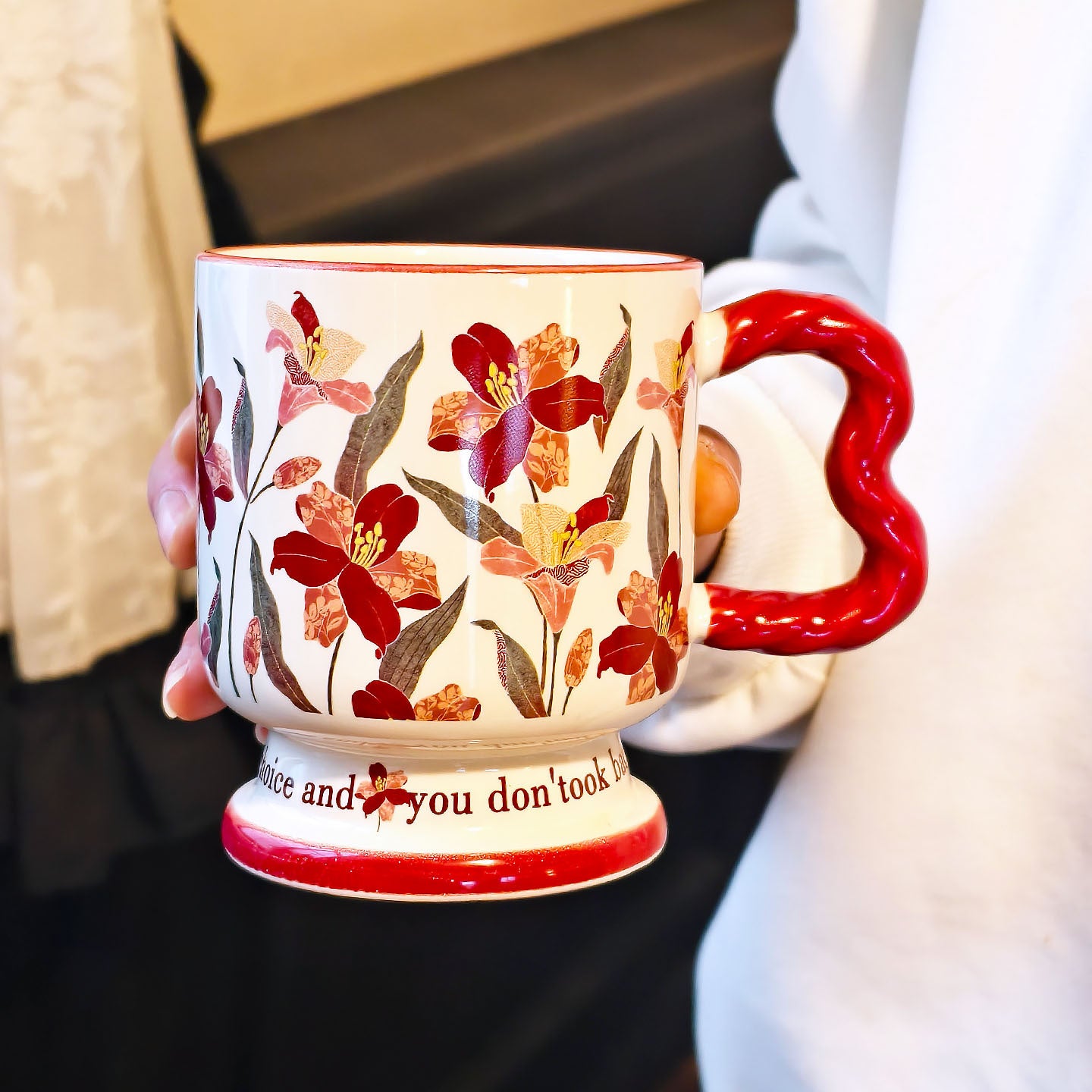 A hand holding a single vintage aesthetic cream ceramic pedestal mug with dark red floral oil painting patterns, featuring a unique red wavy handle and script text on its footed base, over a white surface.