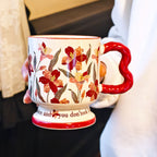 A hand holding a single vintage aesthetic cream ceramic pedestal mug with dark red floral oil painting patterns, featuring a unique red wavy handle and script text on its footed base, over a white surface.