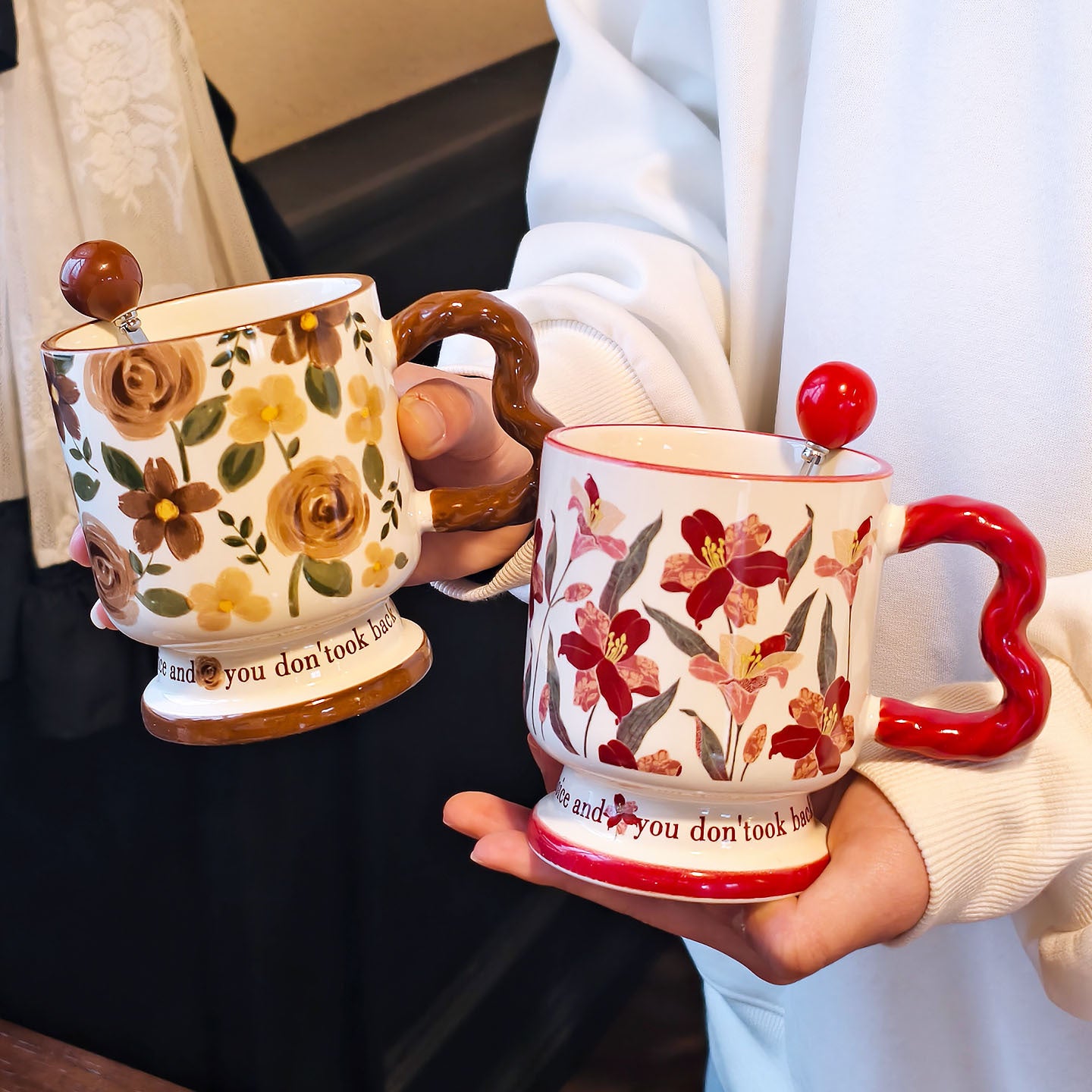 A hand holding two vintage aesthetic ceramic pedestal mugs with floral oil painting patterns and wavy handles. The left mug features brown and yellow roses with a brown wavy handle; the right mug features dark red floral patterns with a red handle. Script text is visible on both bases.