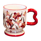 Isolated vintage aesthetic ceramic pedestal mug on a white background, featuring dark red floral oil painting patterns and a red wavy handle. Elegant script text is partially visible on the footed base.