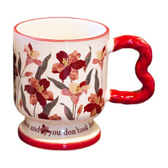 Isolated vintage aesthetic ceramic pedestal mug on a white background, featuring dark red floral oil painting patterns and a red wavy handle. Elegant script text is partially visible on the footed base.