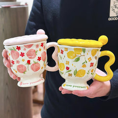 Two aesthetic pedestal mugs with patterned ceramic lids and wavy handles, held by a person. The left mug features peach and red flower patterns with a pink handle and lid; the right mug has lemon and yellow flower patterns with a yellow handle and lid. Both footed bases display the text "love yourself."