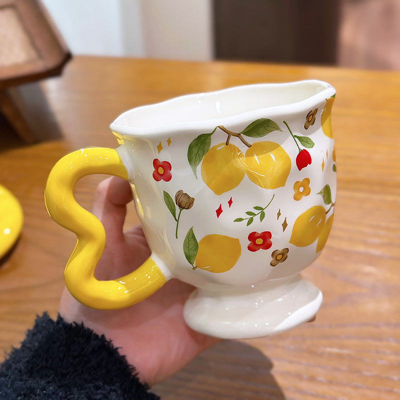 Aesthetic cream ceramic pedestal mug with vibrant yellow lemon and small floral patterns, featuring a unique yellow wavy handle and a delicate scalloped rim, held by a hand over a wooden table.