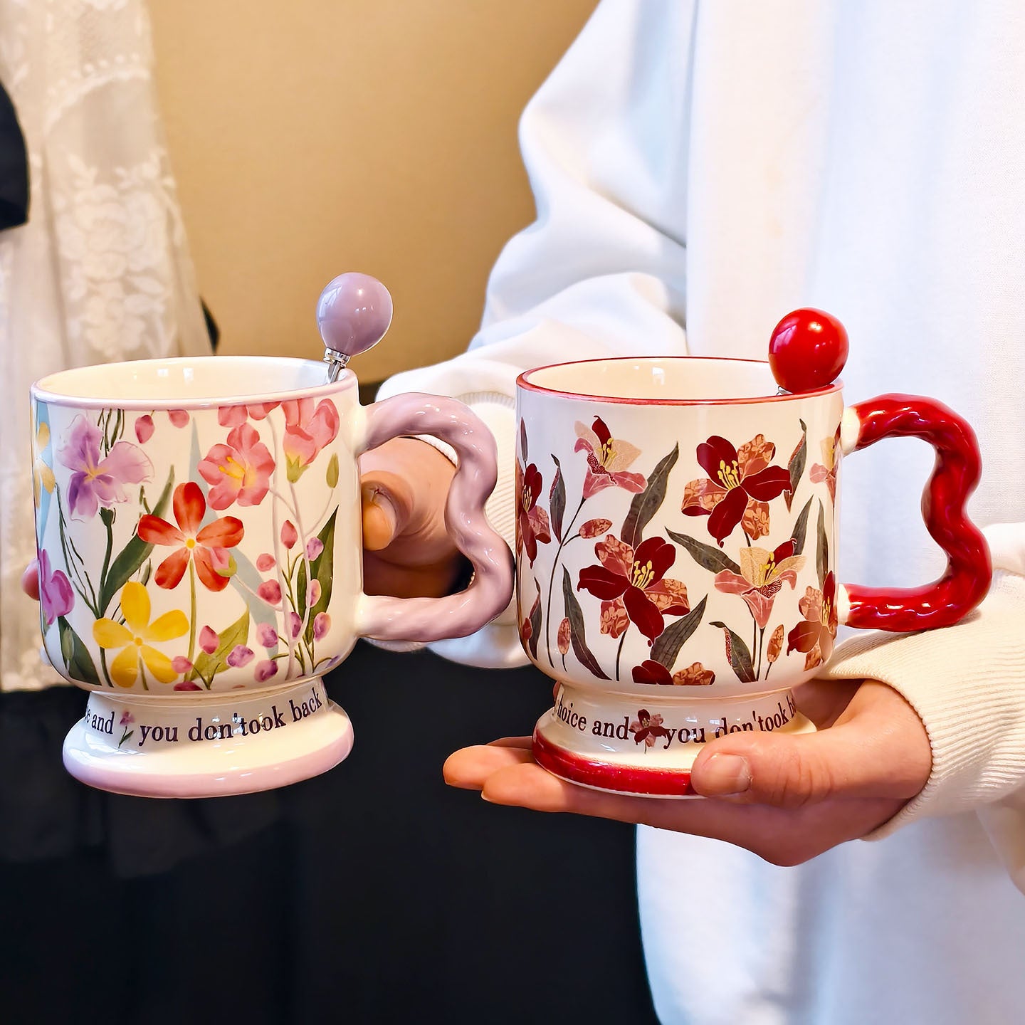 A hand holding two vintage aesthetic ceramic pedestal mugs side-by-side against a soft background. The left mug has purple and pink watercolor flowers with a lavender wavy handle; the right mug has dark red floral patterns with a red handle. Script text is visible on both bases.