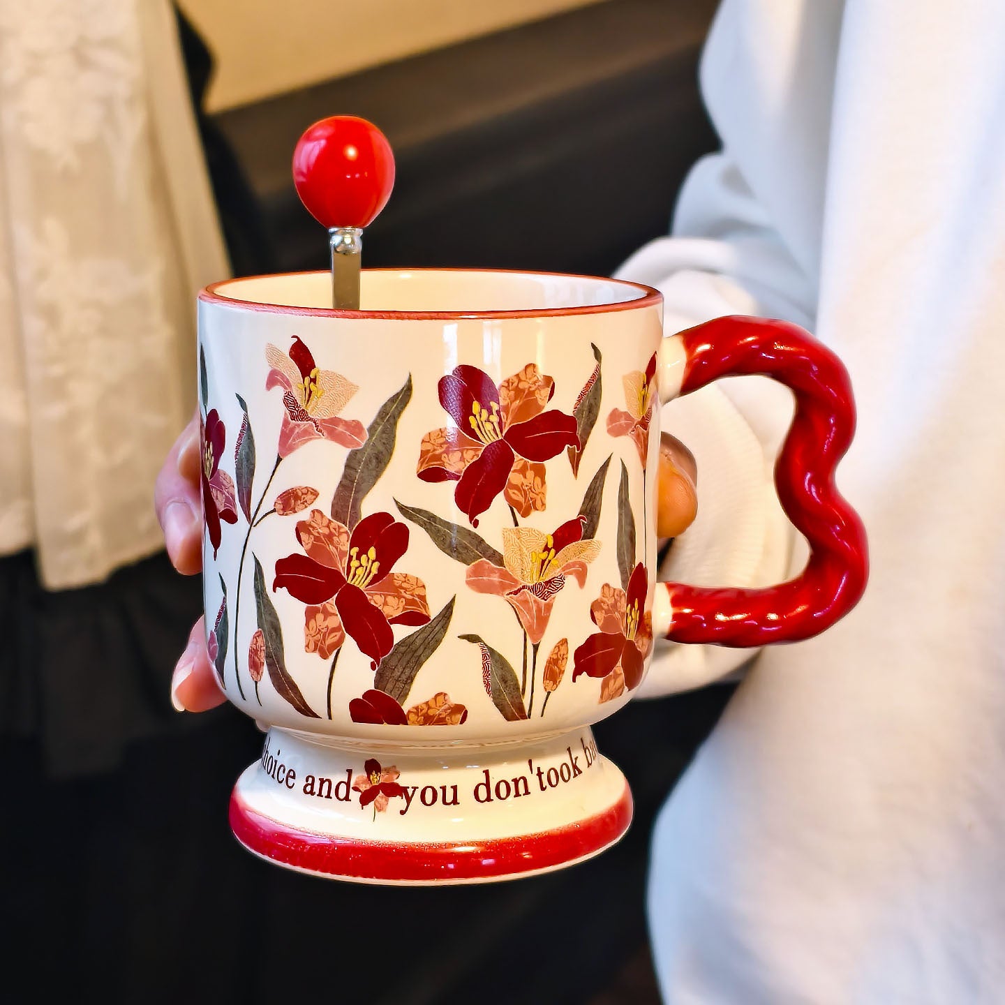 A hand holding a vintage aesthetic cream ceramic pedestal mug with dark red floral patterns and a red wavy handle, with a matching red spherical ball ornament stirring spoon placed inside. Script text is visible on the base.