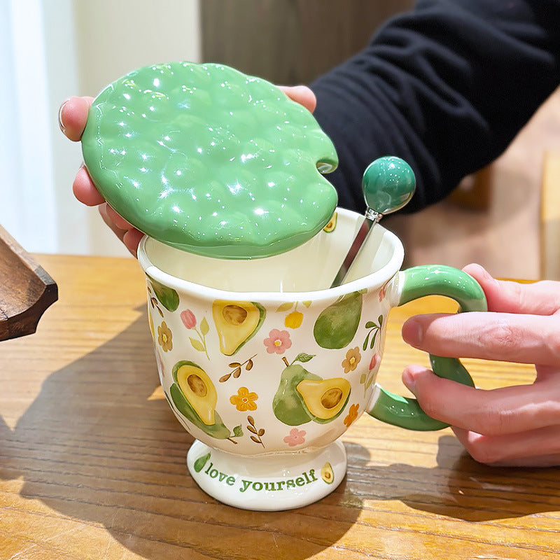 Aesthetic cream ceramic pedestal mug with green avocado and floral patterns, featuring a green wavy handle and 'love yourself' text on the footed base. A hand lifts the matching textured green lid to reveal a stirring spoon with a green ball ornament inside.