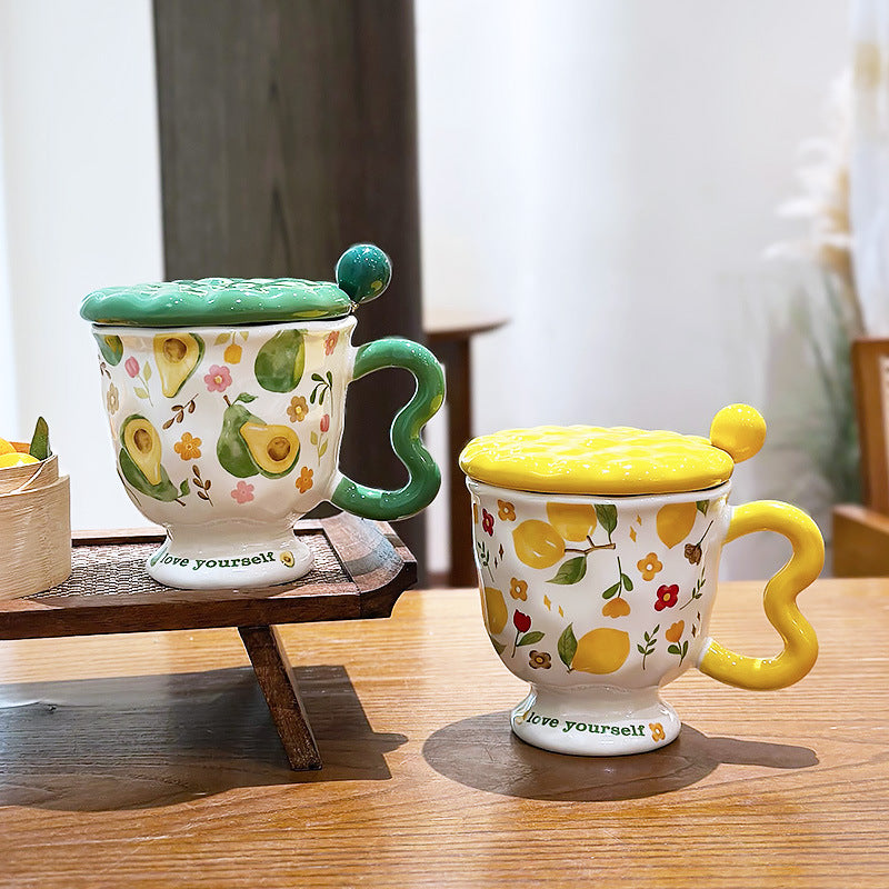 Aesthetic ceramic pedestal mugs with textured lids and wavy handles on a wooden table. The left mug features green avocado and floral patterns with a green handle; the right mug features yellow lemon and floral patterns with a yellow handle. Both footed bases display the text 'love yourself'.