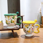 Aesthetic ceramic pedestal mugs with textured lids and wavy handles on a wooden table. The left mug features green avocado and floral patterns with a green handle; the right mug features yellow lemon and floral patterns with a yellow handle. Both footed bases display the text 'love yourself'.