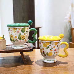 Aesthetic ceramic pedestal mugs with textured lids and wavy handles on a wooden table. The left mug features green avocado and floral patterns with a green handle; the right mug features yellow lemon and floral patterns with a yellow handle. Both footed bases display the text 'love yourself'.