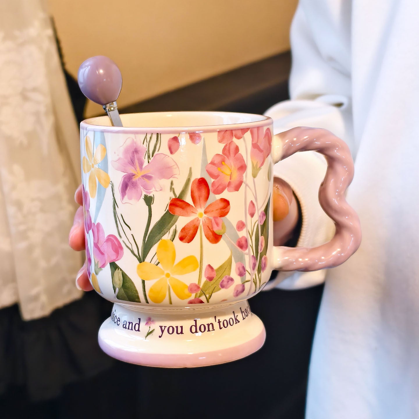 A hand holding a vintage aesthetic cream ceramic pedestal mug with purple and pink floral patterns and a lavender wavy handle, with a matching lavender spherical ball ornament stirring spoon placed inside. Script text is visible on the base.