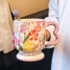 A hand holding a vintage aesthetic cream ceramic pedestal mug with purple and pink floral patterns and a lavender wavy handle, with a matching lavender spherical ball ornament stirring spoon placed inside. Script text is visible on the base.