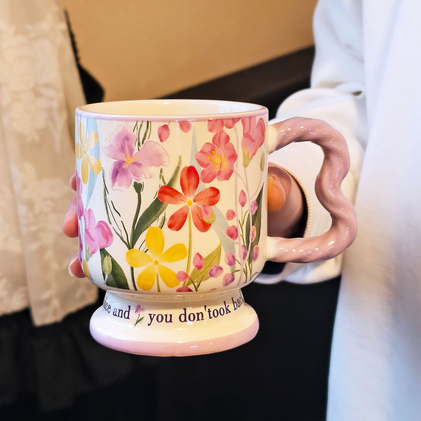 A hand holding a single vintage aesthetic cream ceramic pedestal mug with purple, pink, and yellow watercolor floral patterns, featuring a unique lavender wavy handle and script text on its footed base, over a white surface.